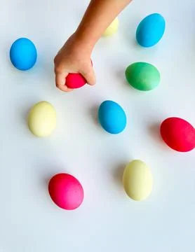 Child's hand picking up blue Easter egg among colorful eggs on white surface Stock Photos