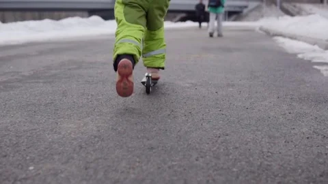 Child's leg in boot pushes off road to ride scooter in winter park, slow motion Stock Footage 167565467