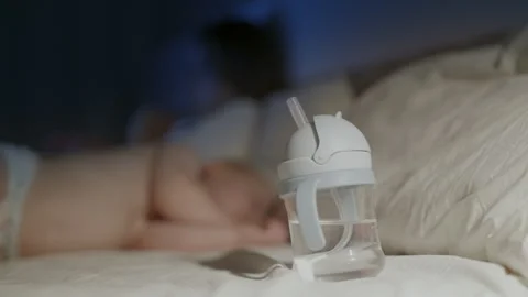 Childs transparent drinking cup with straw placed on bed beside sleeping toddler Stock Footage 341896711