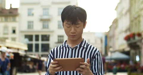 Chinese young man tapping and playing game on tablet device while standing at Stock Footage 125347019