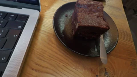 Chocolate cake slice on dark plate beside laptop keyboard on wooden table Stock Footage 337648270