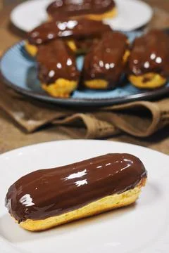 Chocolate Glazed Éclair On Plate With More Pastries In Soft Focus Background Stock Photos