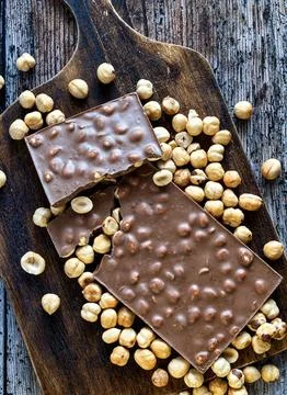 Chocolate tablet with broken hazelnuts on dark wooden board (zenithal view). Stock Photos