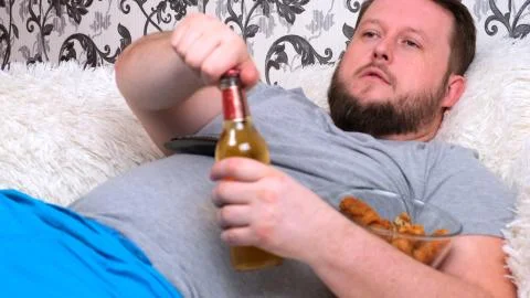 Chubby, fat man with a beard in a gray T-shirt drinks beer and eat fast food Stock Photos