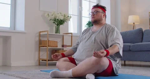 Chubby humorous man practicing seated yoga and mindfulness on mat in cozy home Video stock 329971036