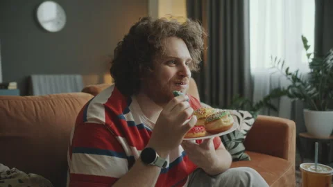 Chubby Man Eating Glazed Donuts and Watching TV in Living Room Stock Footage 303207823