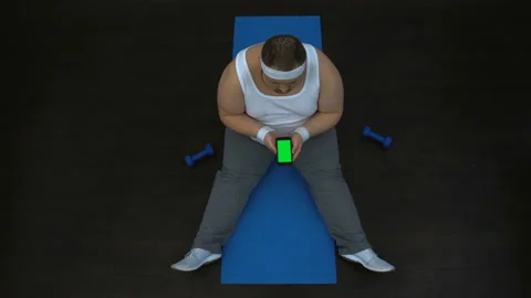 Chubby man using fitness application on smartphone, sitting on mat, green screen Video stock 89518005