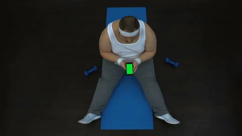 Chubby man using fitness application on smartphone, sitting on mat, green screen Video stock 89533016