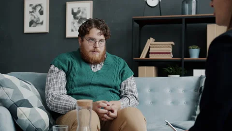 Chubby young man talking to therapist sitting on comfy couch and looking at Video stock 162897680