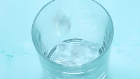 Chunks of ice fall from above into glass beaker on blue background. Close-up. In Stockbeeldmateriaal 247498823