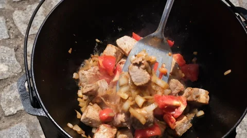 Chunks of meat fried with onions and tomatoes in a cast iron cauldron outdoors Vidéo 346246900