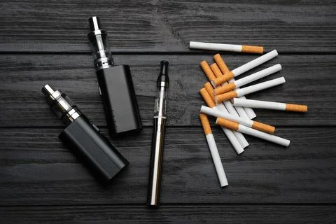 Cigarettes and different vaping devices on black wooden background, flat lay. Stock Photos