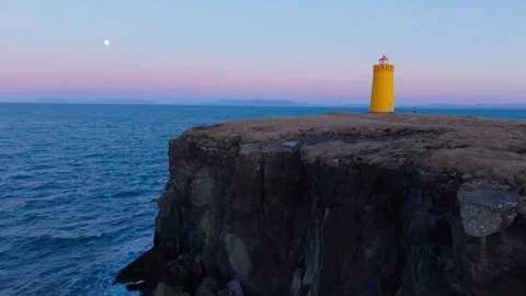 Cinematic drone orbit of a man walking by a yellow lighthouse on basaltic Stock Footage 330895487