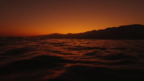 Cinematic Low Angle Dark Ocean Waves at Deep Orange Sunset with Mountain Ridge  Stock Footage 333464403