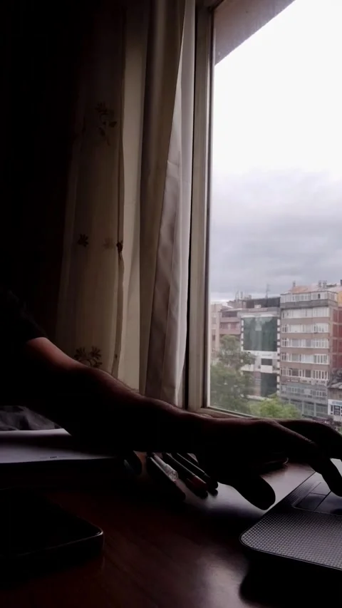 A cinematic low-light shot of a workspace by a window on a rainy day. A hand is Vídeos de archivo 333376252
