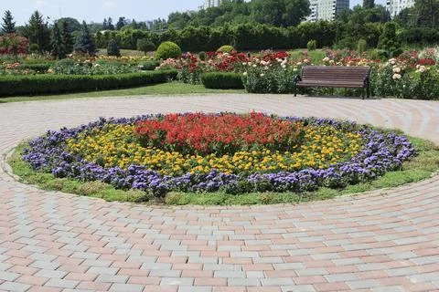 A circular flowerbed features vivid red, yellow, and purple plants, while a.. Stock Photos