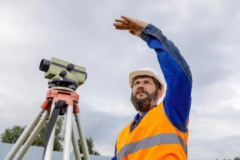 A civil engineer with an optical level waves his hand to control colleagues from Stock Photos