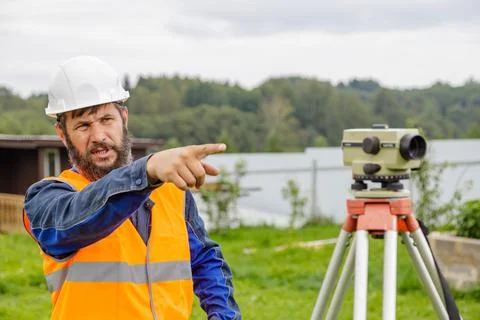 A civil engineer with an optical level waves his hand to control colleagues from Stock Photos