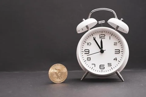 Classic white alarm clock stands next to a bitcoin coin and the time shows 5 库存照片