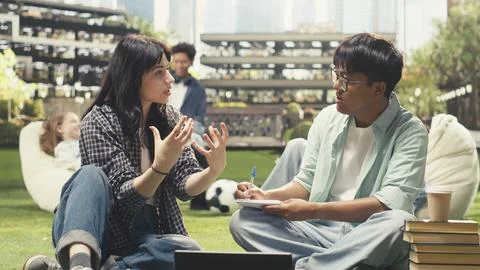 Classmates in study session sitting on grass with laptop placed on stacked books Stock Photos