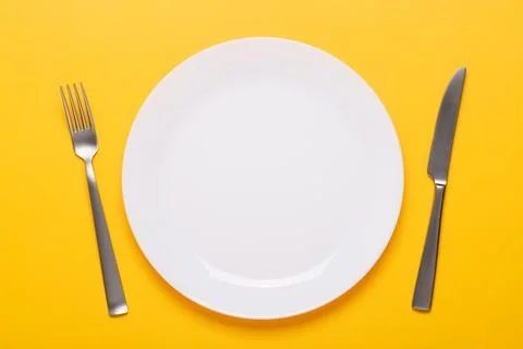 Clean empty white plate with knife and fork on yellow background Stock Photos