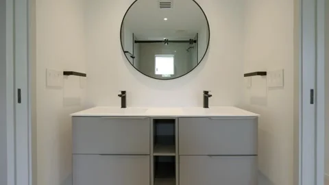 Clean round mirror hanging on wall above white ceramic basin with two taps for Stock Footage 282285502