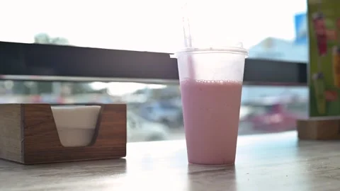 A clear cup filled with a pink beverage sits on a wooden table next to a small Video stock 344526588