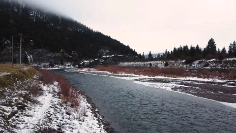 Clear flowing river with snow covered rocky shore and tree covered mountain i Stock Footage 125054183