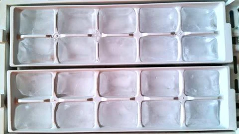 Clear ice cubes sitting inside white plastic kitchen freezer trays Stock Photos