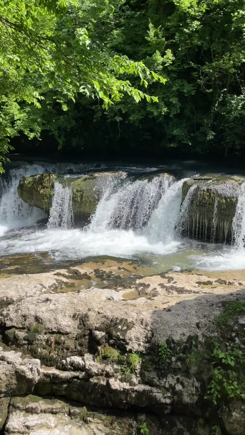 Clear river water pours over a low rocky waterfall beneath lush green fores.. Stock Footage 340559210