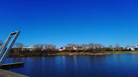 Clear sky over the calm lake with buildings in the background and trees along th 스톡 동영상 332636667