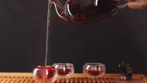 Clear teacup overflows with hot tea drink flowing from a glass teapot held high Stock Footage 207070912