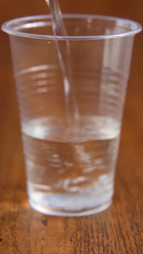 Clear water stream pouring into transparent plastic cup on wooden surface Stock Footage 312648375