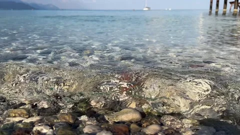 Clear waters waves gently lapping the shoreline, revealing smooth pebbles on a Видео 273782032