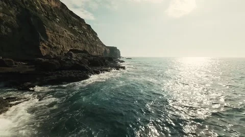 Cliffside waves crash with power, sunlight reflects off sparkling blue water Stock Footage 340226066