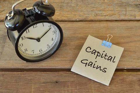 Clock and memo note written with CAPITAL GAINS over wooden background. Stock Photos