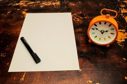 A clock sits on a table next to a piece of paper with a pen on it Stock Photos