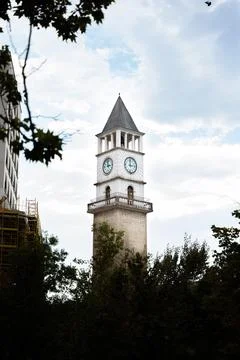Clock in a tower hidden behind trees and bushes in the foreground in Albania Stock Photos