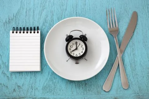 Clock on white plate with fork, knife and notepad, intermittent fasting and w Stock Photos