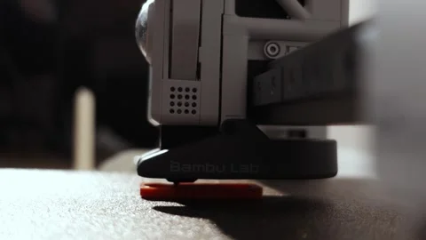 Close-up of a 3D printer nozzle depositing molten plastic onto an orange Видео 339206314