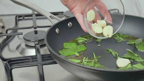Close-up adding garlic cloves into a frying pan with rosemary and sage in oil. Stock Footage 145998702