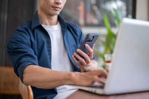 Close up of Asian man hand's using smartphone and typing on laptop at table.. Stock Photos