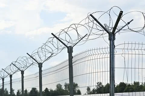 Close up of barbed wire stretched atop steel fence against sky on summer day. Stock Photos