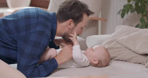 Close of bearded and mustashed father is kneeling by the baby lying on the couch Vidéo 140171399