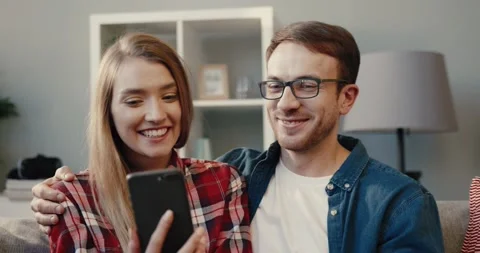 Close up of beautiful young couple sitting at home on couch and looking at funny Stock Footage 148931273