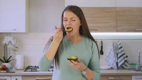 Close up beautiful young woman eats the half of avocado in the kitchen with a Video stock 144751637