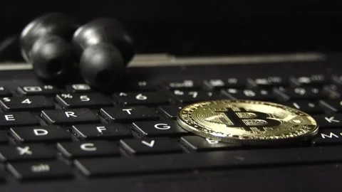 A close-up of a Bitcoin coin falling on a black laptop keyboard with headphones Stock Footage 342256499