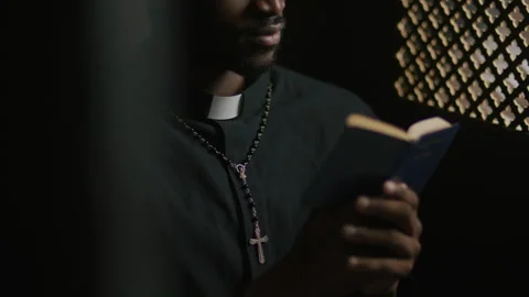 Close-Up of Black Priest Reading Scripture from Bible in Confessional Видео 318340560