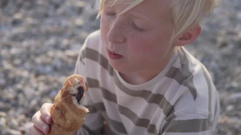 Close-up, a blond boy eating a chocolate croissant on a pebble beach Stock Footage 234643714