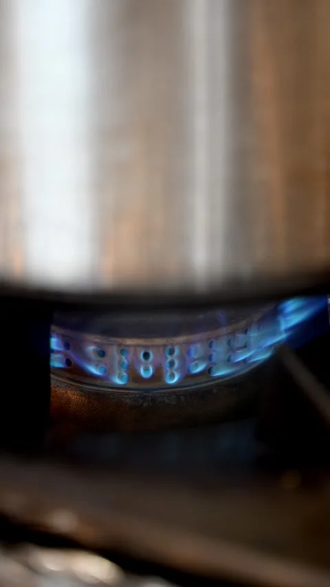 Close-up of a blue natural gas flame burning on a stovetop burner underneat.. Видео 338053655